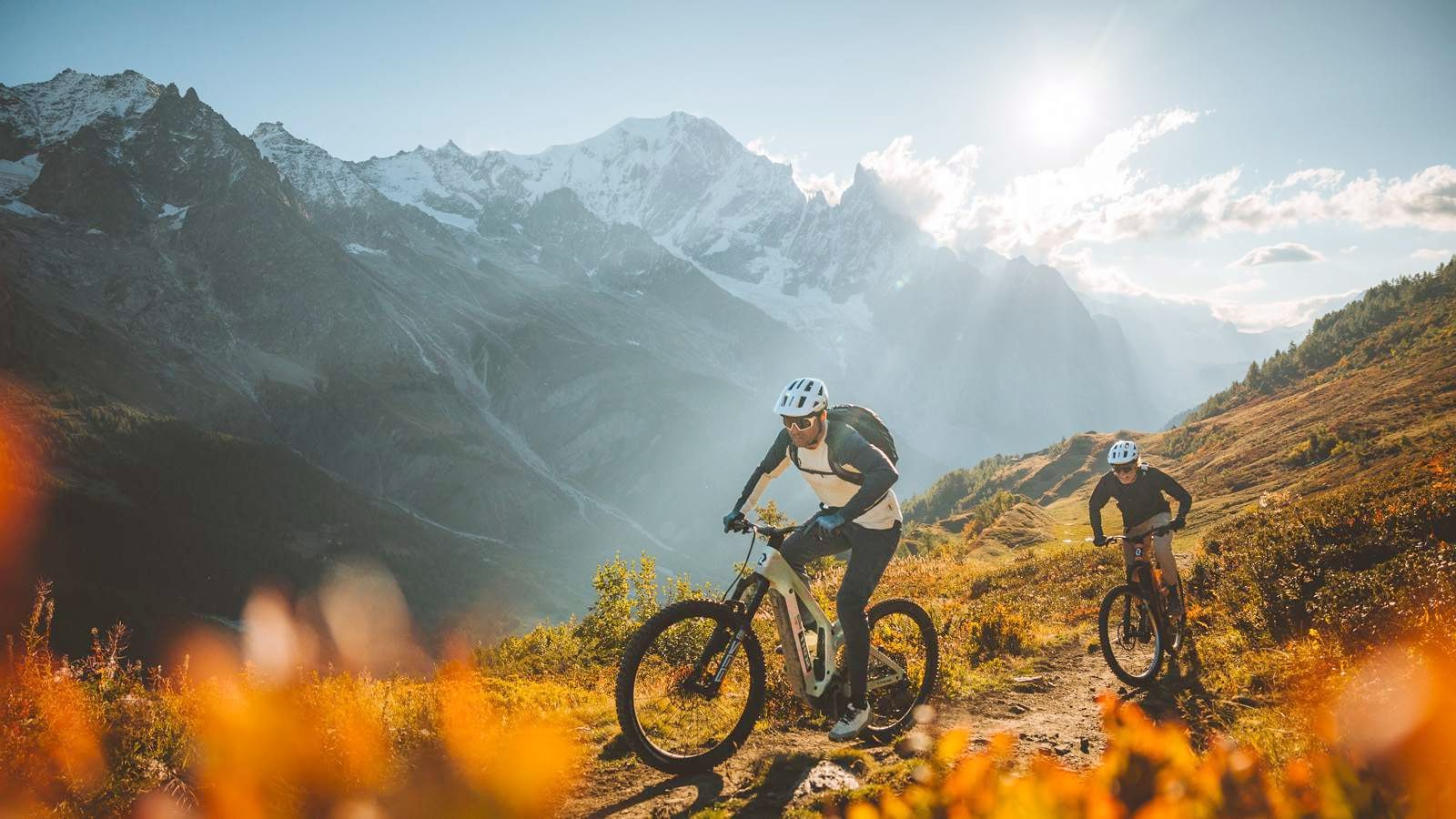 Two riders climbing an alpine trail in autumn, snow peaks behind